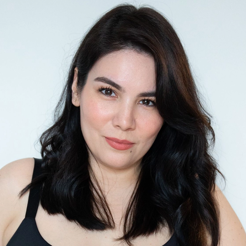 A woman with medium-length wavy black hair, fair skin, and dark eyes looks at the camera. She’s wearing a black sleeveless top, has natural makeup with peach lipstick, and is standing against a plain light background. She has a calm, confident expression.