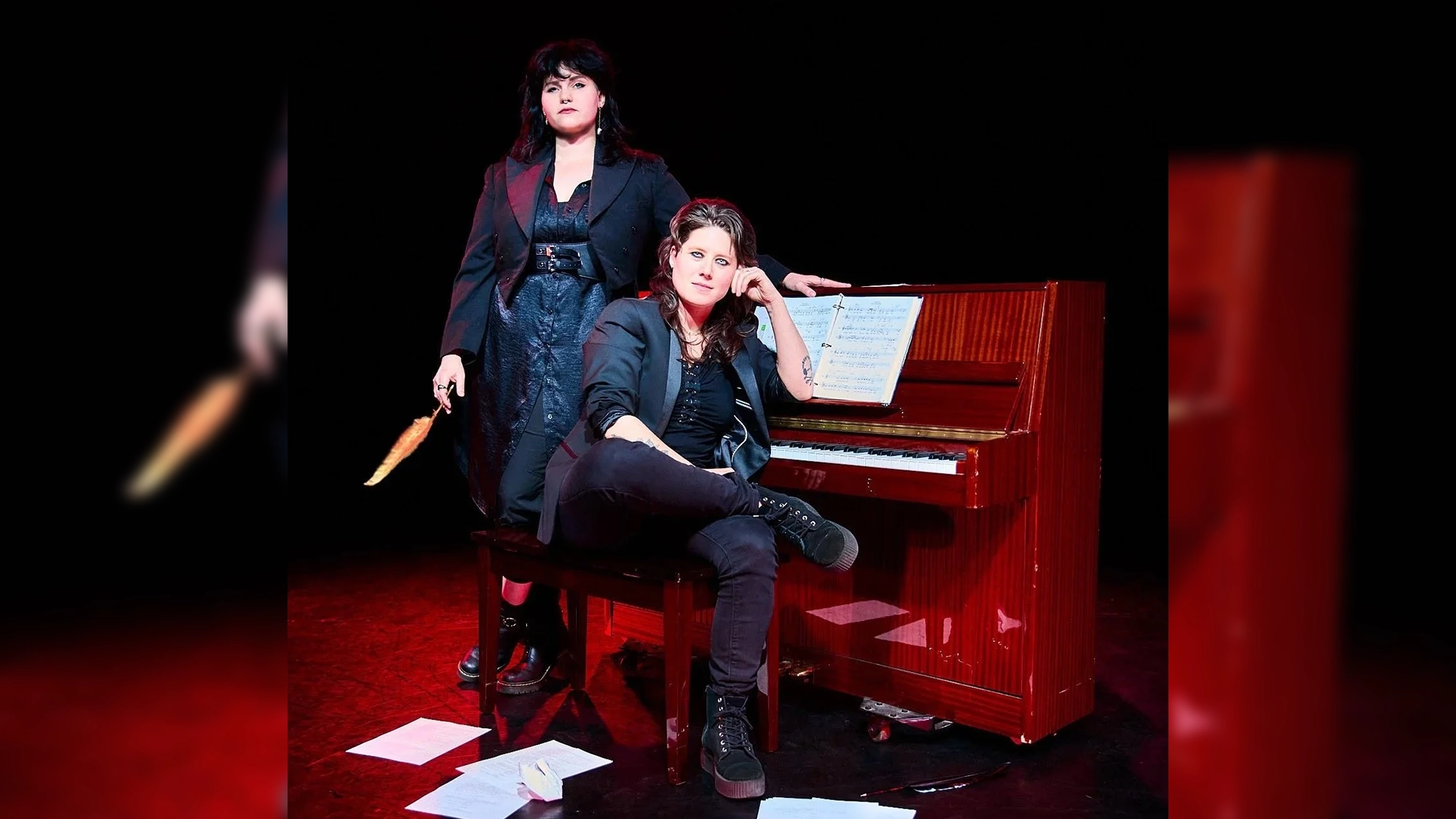 Two women pose by a piano on a dark stage, creating an evocative scene reminiscent of a queer musical. One stands holding a feather, dressed in a black dress and coat, while the other—perhaps evoking Atalanta from Lucier&Rose—sits on the piano bench in black clothing, looking at the camera. Sheet music and papers are scattered across the floor, adding to the.
