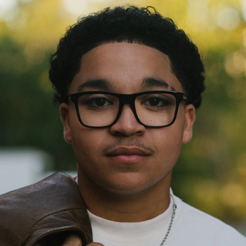 Zeek Atkinson, a young man with short curly hair and glasses, looks at the camera. He wears a white shirt and holds a brown jacket over his shoulder. The softly blurred greenery in the background gives a warm, outdoor feel.