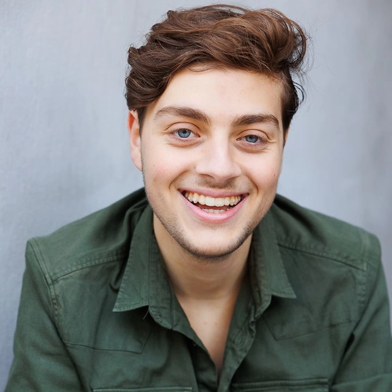 A young man with wavy brown hair and blue eyes smiles warmly. Kristian Babian wears a dark green button-up shirt and sits in front of a plain light gray background. His expression is cheerful and friendly, and he faces the camera directly.