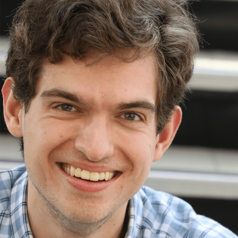 A young man with wavy brown hair and light skin, Sean Barnett, smiles at the camera. He is wearing a light blue plaid shirt. The background is blurred, featuring horizontal black and white lines, possibly benches or bleachers.