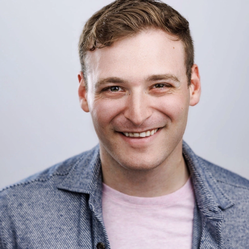 A young man with short, light brown hair smiles warmly at the camera. He is wearing a light pink t-shirt under a blue textured jacket. The background is plain and light, giving a clean, professional appearance to the headshot.