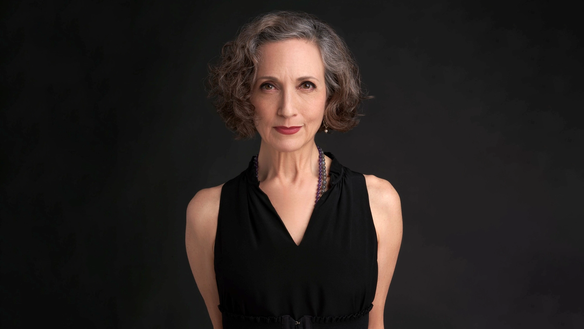 A middle-aged woman with short, curly gray hair wears a sleeveless black dress and a dark beaded necklace. She stands against a plain dark background, looking confidently at the camera with a slight smile.