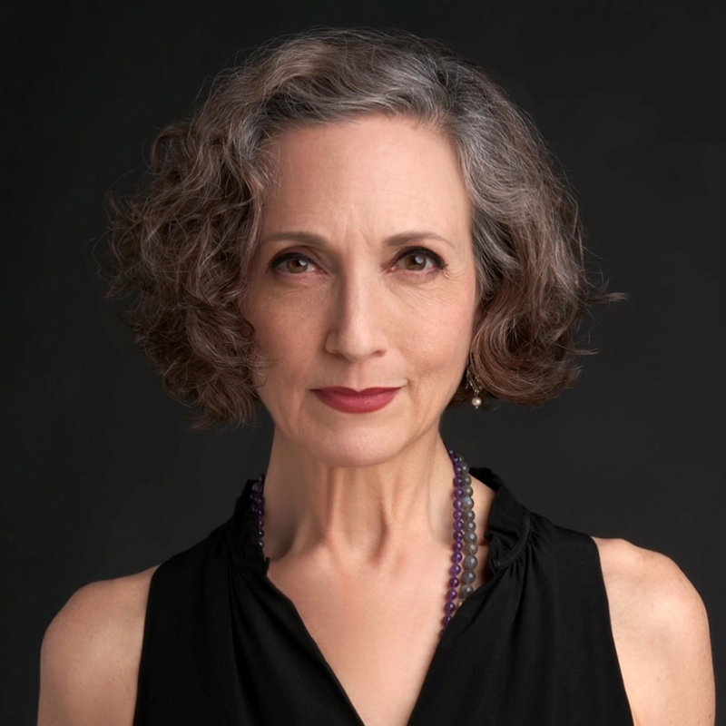 A middle-aged woman with curly gray hair and pearl earrings wears a sleeveless black top and a beaded necklace. She looks directly at the camera with a calm, confident expression, set against a plain dark background.