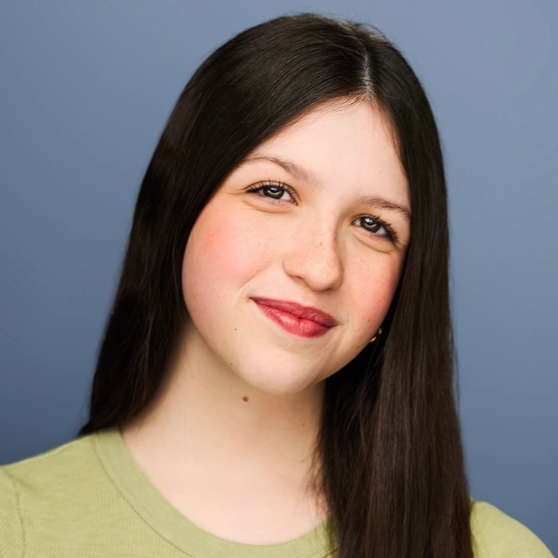 A young person with long straight dark hair and fair skin smiles gently at the camera. They wear a light green shirt and have a small beauty mark on their chin. The background is a smooth, muted blue, perfectly complementing the calm presence of Gaël Mouton-Sady.
