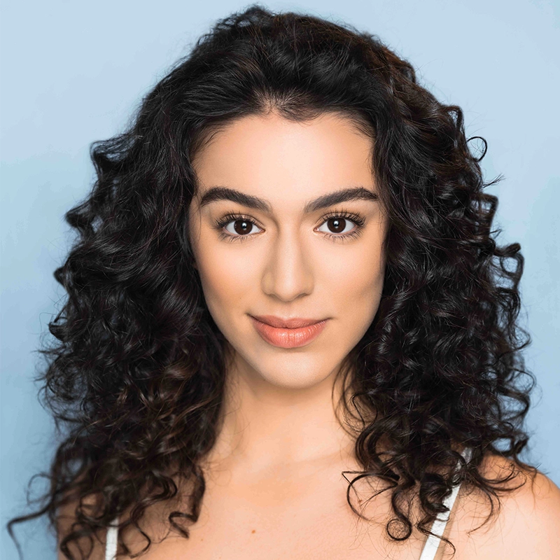 A woman with long, dark, curly hair, brown eyes, and smooth skin gazes at the camera. She wears natural makeup and a white top, set against a light blue background. Her expression is calm and confident, embodying the poise and grace of Gabrielle Farley.
