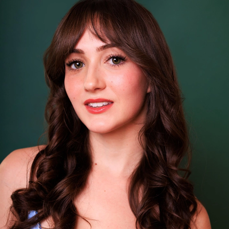 A young woman with long, wavy brown hair and bangs poses against a dark green background. She has light skin, soft makeup with pink lipstick, and wears a light blue top. Bella Birdsley gazes slightly off-camera, smiling gently.