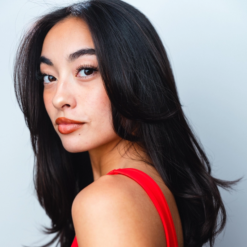 A woman with long, wavy dark hair looks over her shoulder at the camera. She has clear skin, winged eyeliner, and peach lipstick. She wears a sleeveless red top. The background is plain and softly lit.