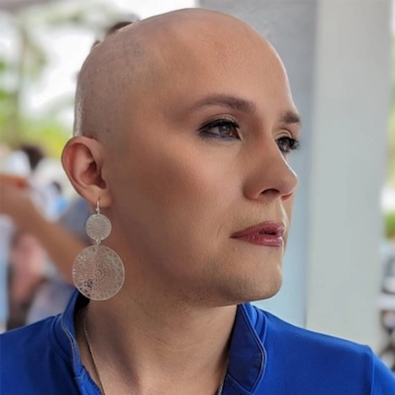 Aaron Valentine, with a shaved head, wearing a blue top and large, circular dangling earrings, looks thoughtfully to the side. Their makeup is subtle, with defined brows and eyes. The softly blurred background suggests an outdoor setting.