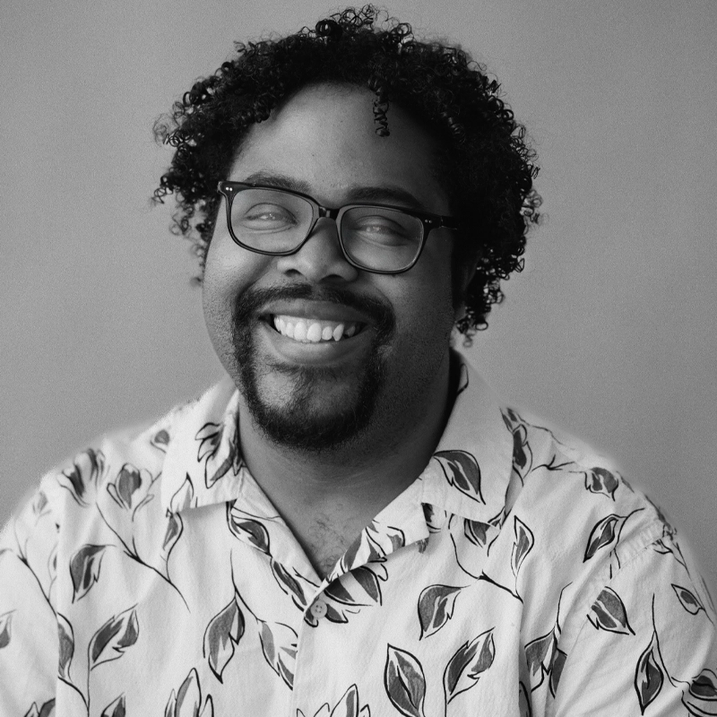 Brandon Bogle, a man with curly hair, glasses, and a goatee, is smiling broadly. He wears a light-colored shirt with a bold leafy pattern. The background is plain and softly lit in this black and white photo.