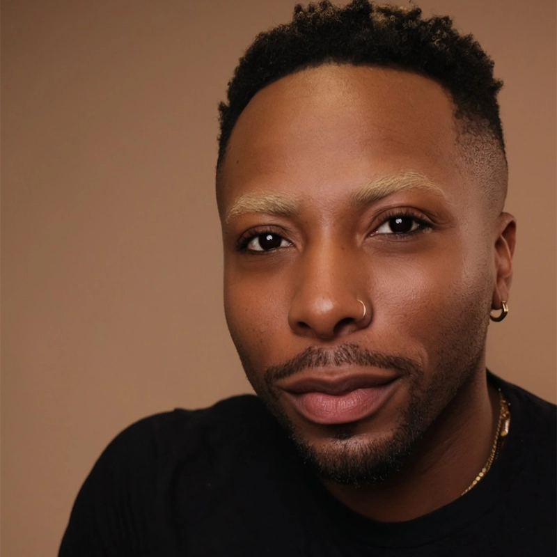 A person with short curly hair, bleached eyebrows, a nose ring, and an earring smiles slightly. They wear a black shirt and a gold chain. The background is a plain, warm beige color, and the lighting is soft, highlighting their facial features—a portrait that exudes effortless style and confidence reminiscent of Olutayo Bosede.