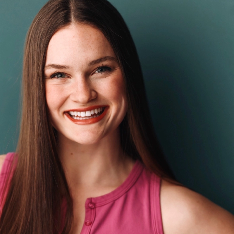 A young woman with long straight brown hair smiles brightly. She is wearing a sleeveless pink top and dark red lipstick. The background is a solid teal color. Her head is slightly tilted, and she looks directly at the camera.