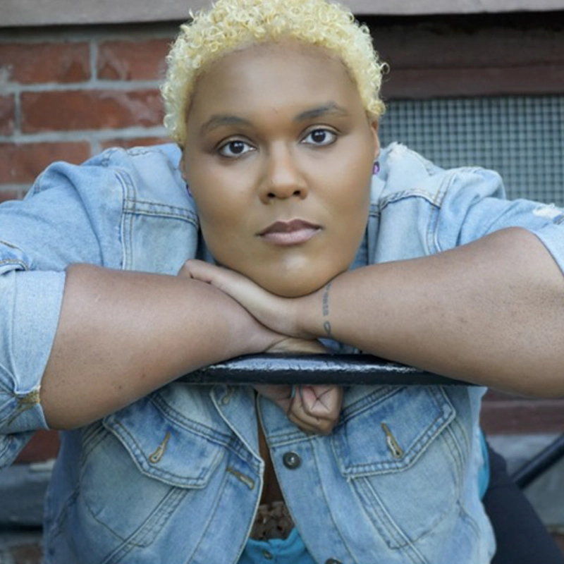 Amara Janae Brady, with short, curly blond hair and brown skin, rests their chin on folded arms over a black metal railing. Wearing a light blue denim jacket, they gaze calmly and seriously at the camera. A brick wall is in the background.