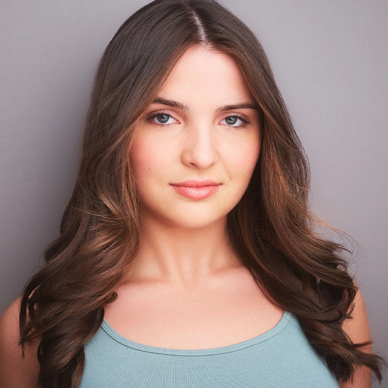 A young woman with long, wavy brown hair and fair skin faces the camera, wearing a light blue sleeveless top. Dru Bramblett has blue eyes and a gentle, confident expression against a simple gray background.
