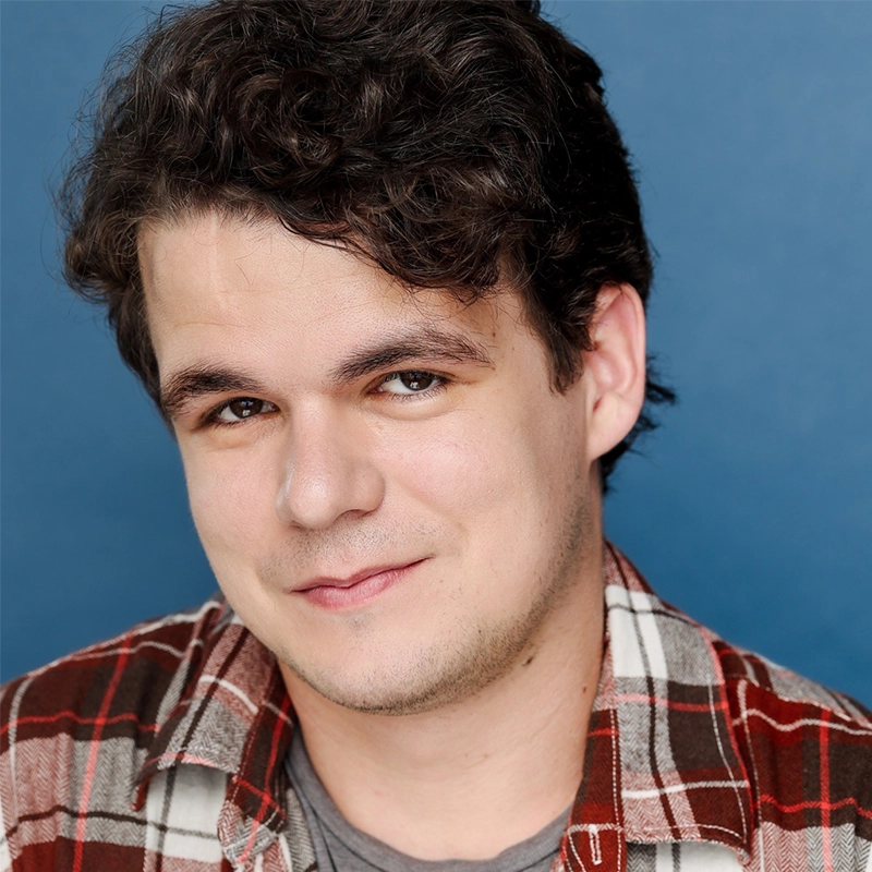 A young man with short dark curly hair smiles slightly. He wears a red and brown plaid shirt over a gray t-shirt. The background is solid blue. Casey Keeler faces the camera in a relaxed pose, and his expression is warm and approachable.