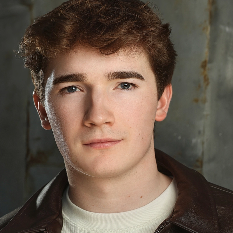 Young man, Saylor Tucker, with short, wavy brown hair and fair skin looks directly at the camera. He wears a brown leather jacket over a cream shirt. The background is gray with slight rust marks, giving an industrial feel. His expression is neutral and calm.