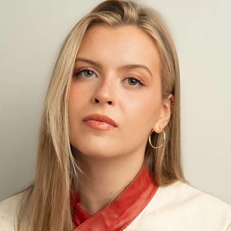 A young woman with long blonde hair wears gold hoop earrings, subtle makeup, and a neutral expression. She has fair skin and blue eyes. Gabrielle Farley is dressed in a cream-colored top with a red leather collar against a plain light background.