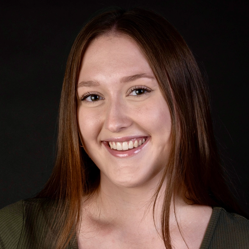 A young woman with straight brown hair smiles at the camera. She has fair skin, light makeup, and wears a dark green top. The solid black background, characteristic of Phillip Wong’s portrait style, highlights her face. Her expression is friendly and approachable.