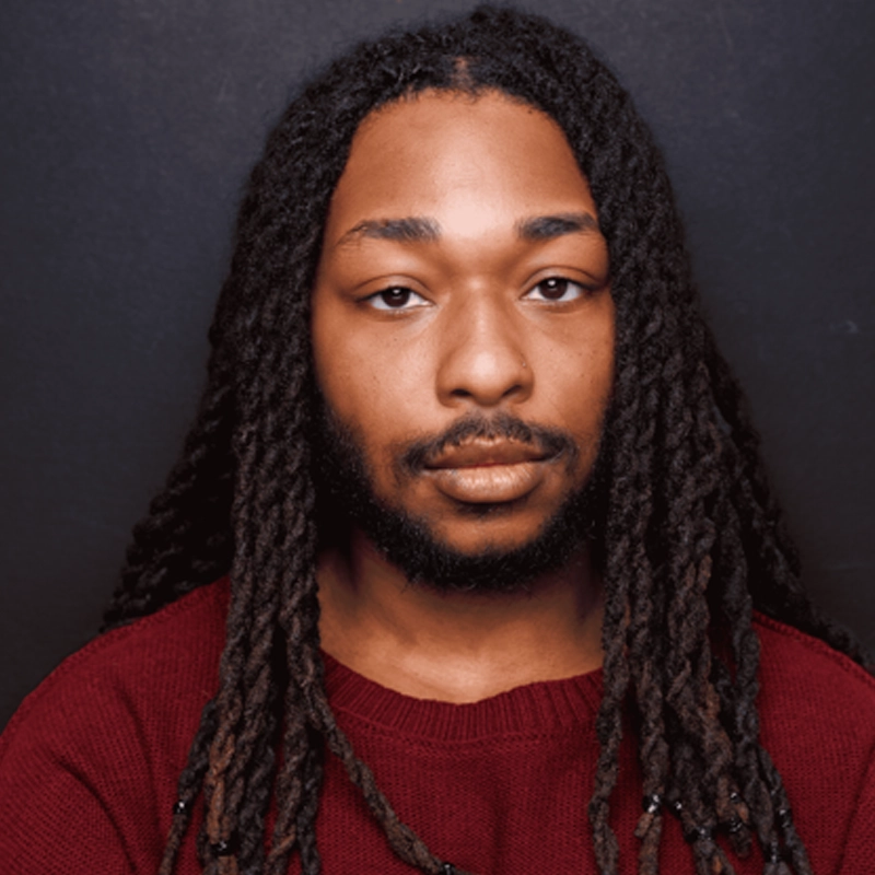 A person with long, dark, and slightly brown-tinted dreadlocks wears a maroon sweater and looks directly at the camera with a neutral expression. The background is plain and dark, creating a strong contrast with their attire and hair.