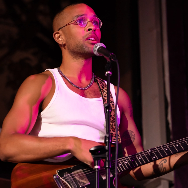 A person with short hair, glasses, and hoop earrings plays an electric guitar and sings into a microphone. Xander Browne wears a white tank top, chain necklace, and a leopard print guitar strap under pink stage lighting.