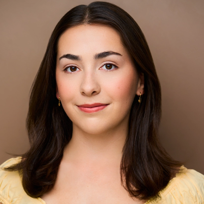Ainsley Burns, a young woman with straight, dark brown hair parted in the middle, wears small gold earrings and a light yellow top as she smiles softly against a plain brown background. Her makeup is natural, and she appears calm and confident.