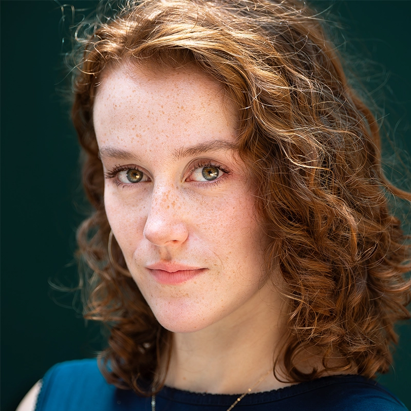 A young woman with fair skin, green eyes, and curly red hair looks directly at the camera. She has light freckles, a slight smile, and wears a navy blue top. The background is a solid dark green, emphasizing her features in a style reminiscent of Gaël Mouton-Sady.