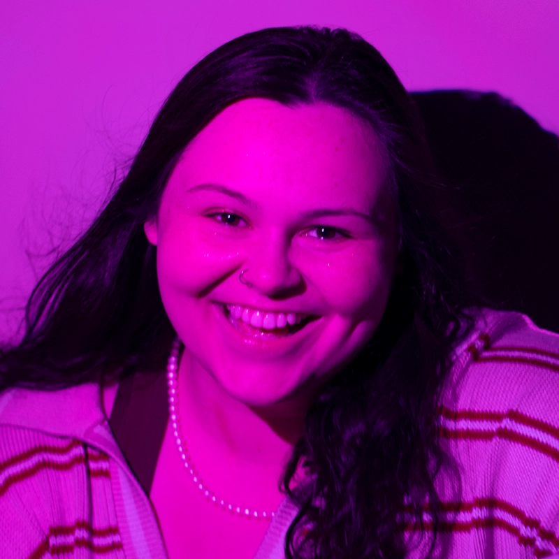 A young woman with long dark hair smiles at the camera. She wears a striped sweater and a pearl necklace. The image is tinted bright pink, giving a vibrant effect. The background is plain, and her dimples are visible as she smiles—capturing the confident energy of 