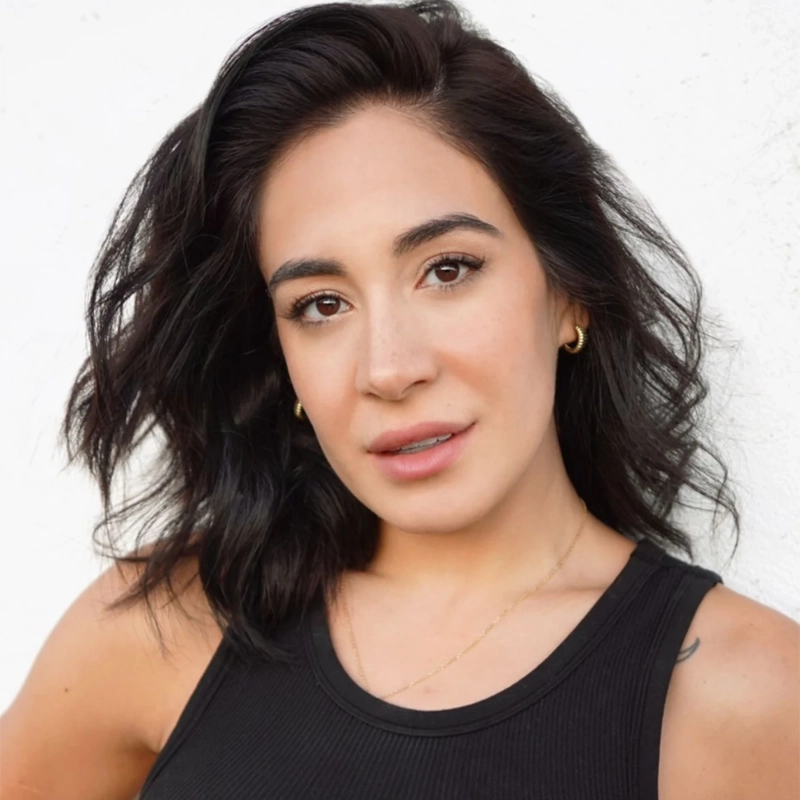 A woman with wavy dark hair, light makeup, and gold earrings wears a black tank top and a delicate gold necklace. She stands against a plain white background, looking directly at the camera with a neutral, relaxed expression—her style reminiscent of the understated elegance often associated with Gabriela Carrillo.