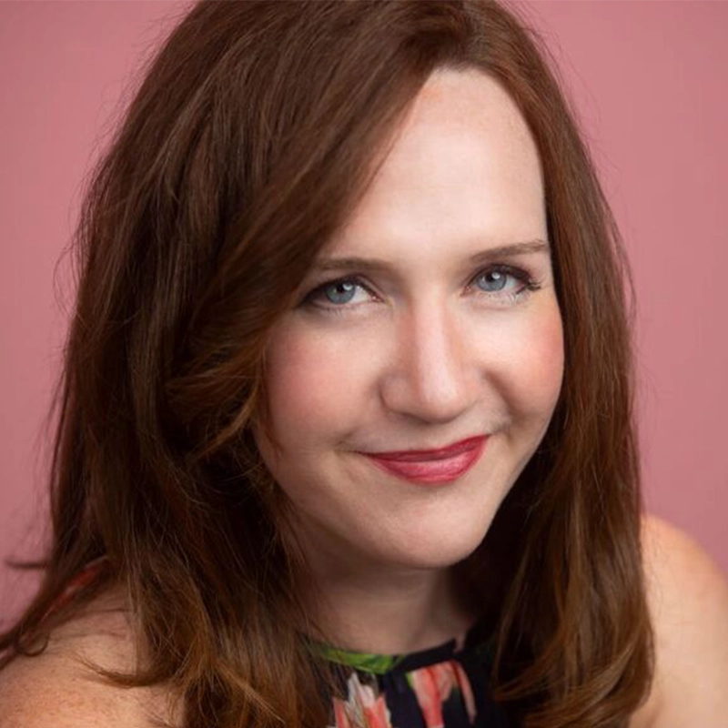 Casey Keeler, a woman with long auburn hair, smiles softly at the camera. She is wearing a sleeveless floral top and red lipstick. The background is a plain pink color. Her blue eyes and light skin are visible in the close-up portrait.