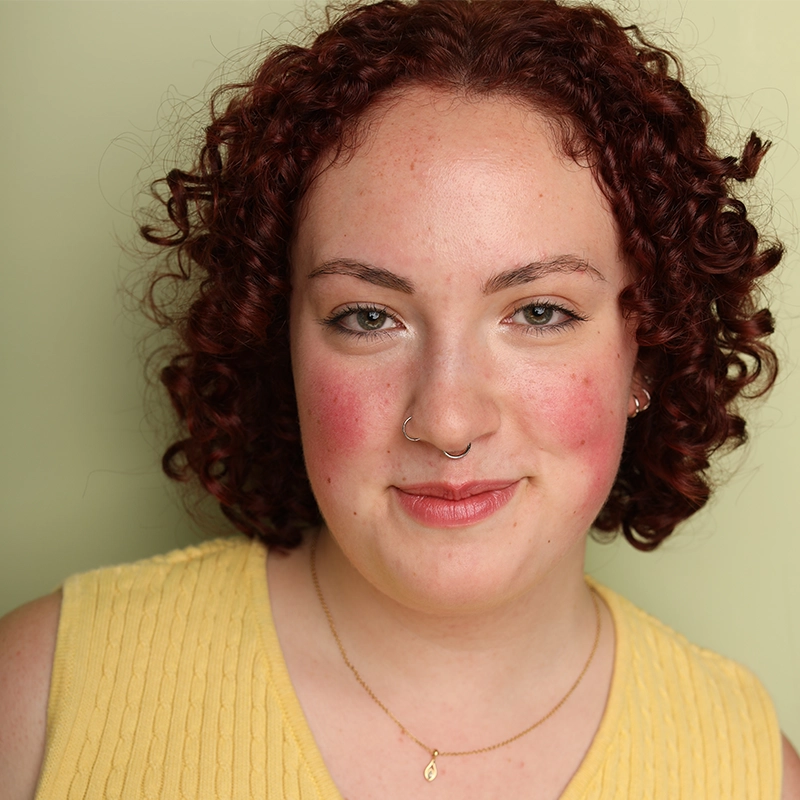 Jayline Ross, a young person with fair skin, curly auburn hair, and flushed cheeks, smiles slightly. They wear a yellow sleeveless top, a gold nose ring, and a delicate gold necklace while standing against a pale green background.