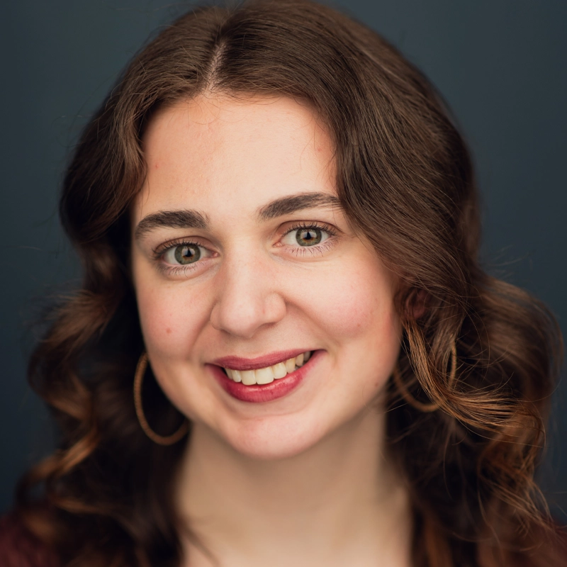 Mackenzie Cavagnaro, a young woman with wavy brown hair and green eyes, smiles at the camera. She wears large hoop earrings and a burgundy top. The background is a plain, dark blue. Her skin is fair, and her makeup is natural, highlighting her lips and eyes.