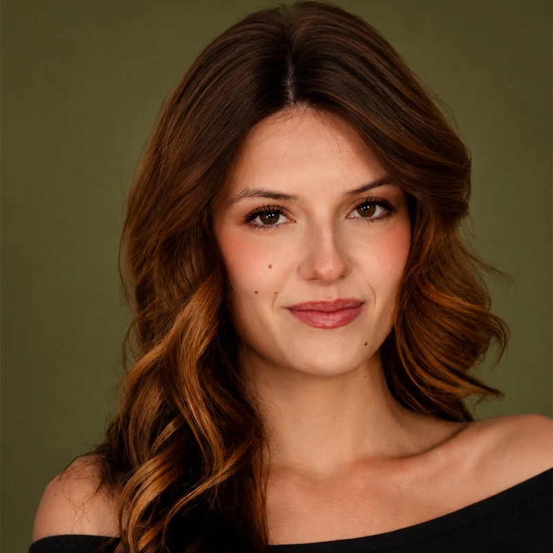 Margaux Champon, a woman with long wavy brown hair and light skin, wearing an off-the-shoulder black top, poses in front of a plain olive green background, smiling softly with neutral makeup and a small beauty mark on her cheek.