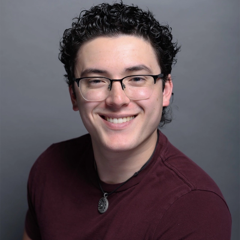 A young person with short, curly dark hair and glasses smiles at the camera. They wear a maroon t-shirt and a black cord necklace with a circular pendant. The background is a smooth, neutral gray.