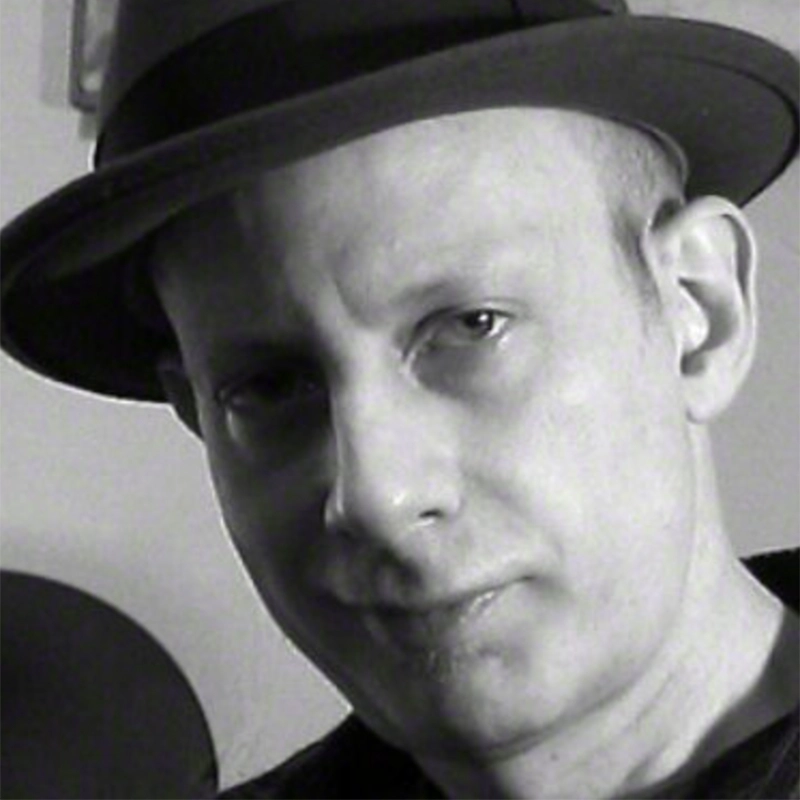 Black-and-white photo of a light-skinned person wearing a dark fedora hat, slightly tilted, looking at the camera with a neutral expression. The background is plain and out of focus. Only the head and part of the shoulders are visible, capturing a timeless portrait style reminiscent of Daniel A Weiss.