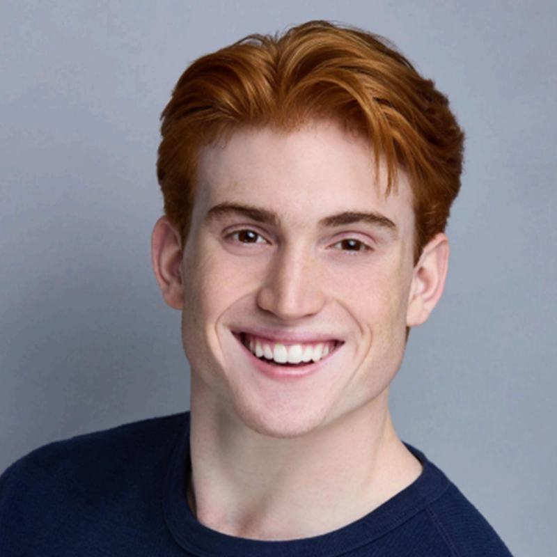 A young man with short, wavy red hair smiles broadly. He wears a dark blue crew-neck shirt and is posed against a simple, light gray Surfac background. His teeth are visible, and he appears cheerful and friendly.