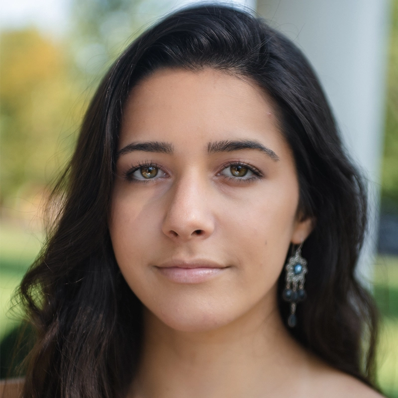 A young woman with long dark hair, wearing ornate drop earrings, gazes softly at the camera outdoors. Her expression is calm, with soft makeup. The background is blurred, showing greenery and a white column. Sunlight highlights her face, capturing Alexandra Zografos in a serene moment.