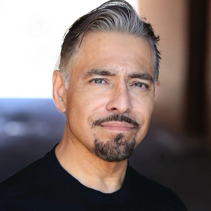 Middle-aged man with short, slicked-back salt-and-pepper hair, light skin, and a trimmed goatee, wearing a black crew-neck shirt. He has a slight smile and is standing in a softly lit area with a blurred brown and blue background.