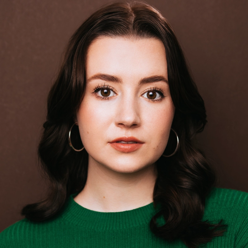 A young woman with wavy, shoulder-length dark hair wears gold hoop earrings and a green sweater. She has light skin, natural makeup, and looks directly at the camera with a neutral expression against a brown background—a timeless portrait reminiscent of Bridget DeVine’s understated elegance.