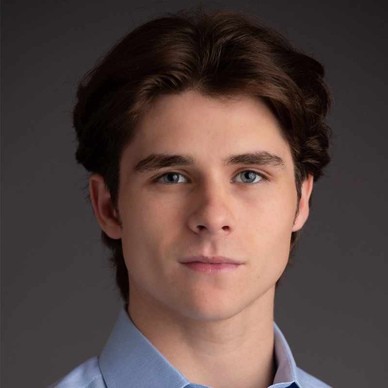 Gaël Mouton-Sady, a young person with fair skin, medium-length brown hair, and blue eyes, looks directly at the camera. They wear a light blue collared shirt and have a neutral expression. The background is dark and plain.
