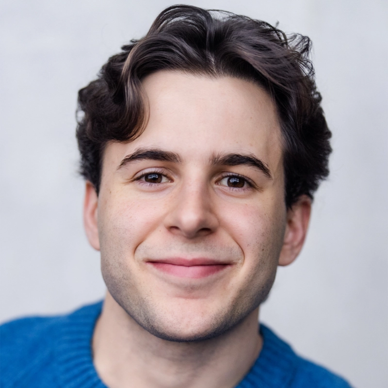 A young man with wavy brown hair and fair skin smiles at the camera. He is wearing a bright blue sweater. The background is plain and light, keeping the focus on his face—perfectly highlighting Jeremy Dermovsesian’s friendly expression.