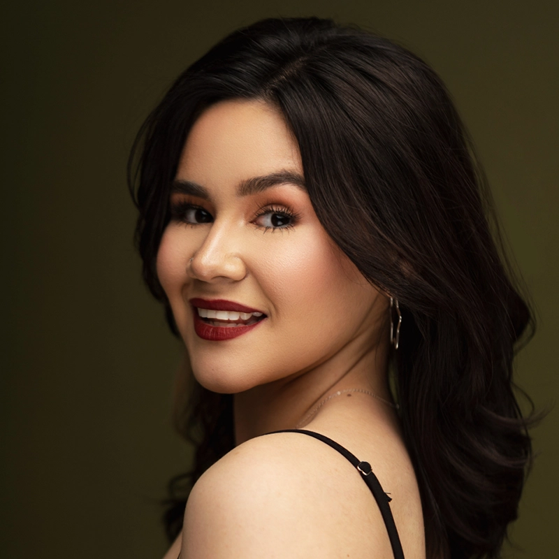 A woman with long, wavy dark hair smiles over her shoulder toward the camera. She wears bold red lipstick, dark eye makeup, a black spaghetti strap top, and hoop earrings. The background is a plain olive green. Her skin appears smooth and glowing.