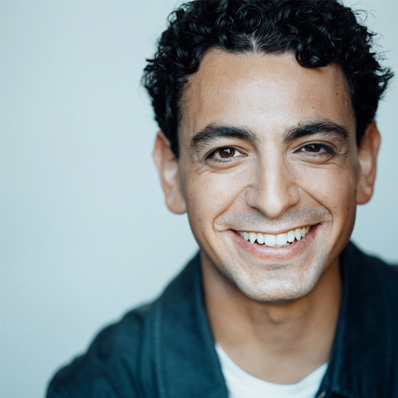 A young man with curly dark hair smiles warmly at the camera. He wears a dark jacket over a white shirt. The background is a soft, light blue. His expression is friendly and welcoming, and the image is well-lit with a close-up focus on his face.