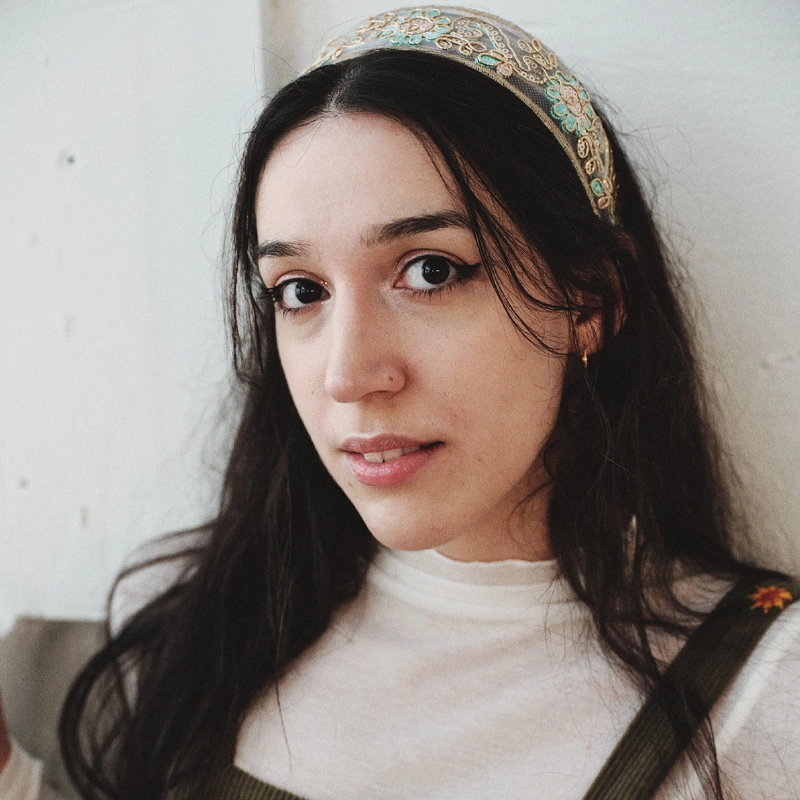 A young woman with long dark hair, reminiscent of Elené Dolidze, wears a decorative headband with gold and turquoise accents. She has light skin, soft makeup, and is dressed in a white top with olive-green overalls, gazing softly at the camera against a light background.