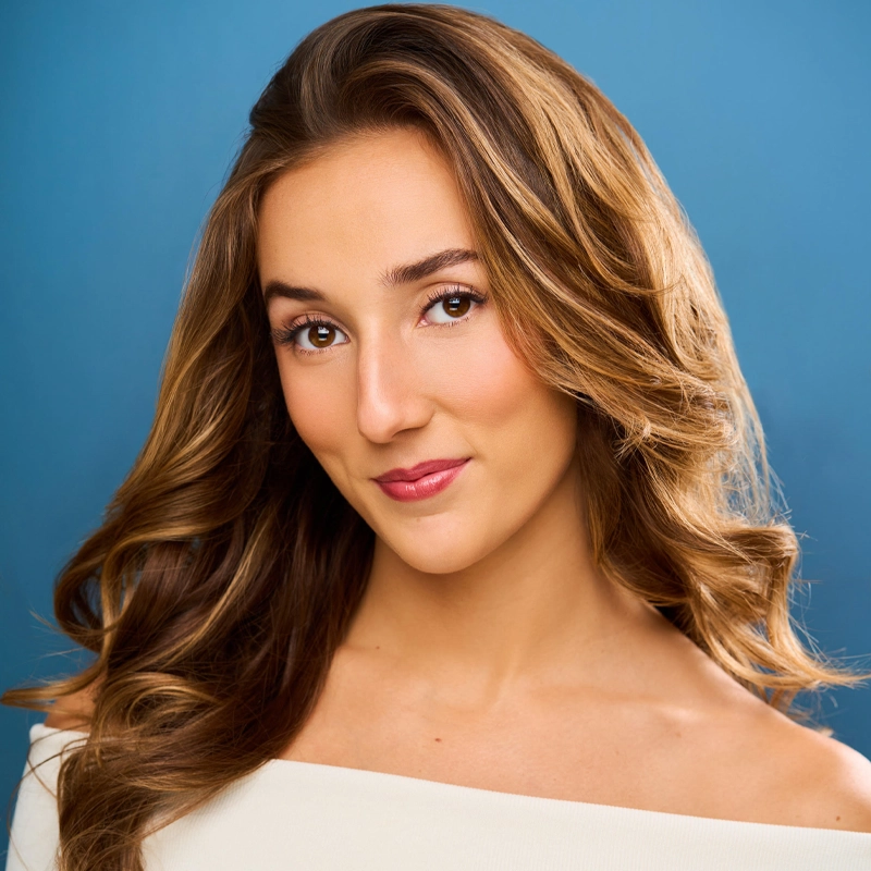 A young woman with long wavy brown hair smiles softly at the camera. She has light skin, brown eyes, and is wearing a white off-shoulder top. The background is solid blue. Her makeup is natural, with soft pink lipstick and subtle blush.