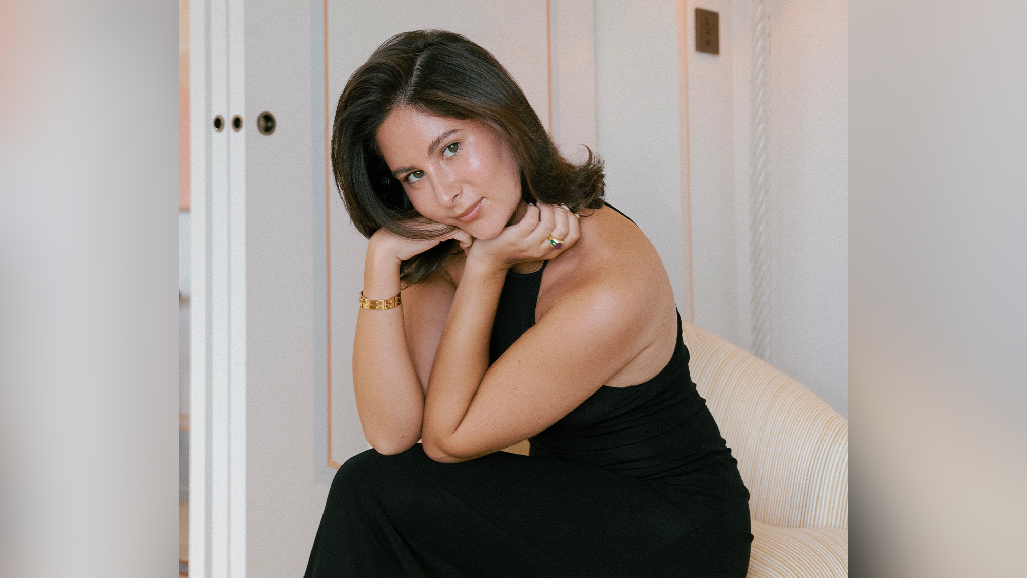 A woman with short dark hair and green eyes sits on a cream chair indoors, wearing a sleeveless black dress, gold bracelet, and ring. She rests her chin on her hands and looks directly at the camera, evoking the poised elegance of Eden Plepler. The neutral background features light paneling, creating a serene setting reminiscent of an intimate Holiday Songbook performance.