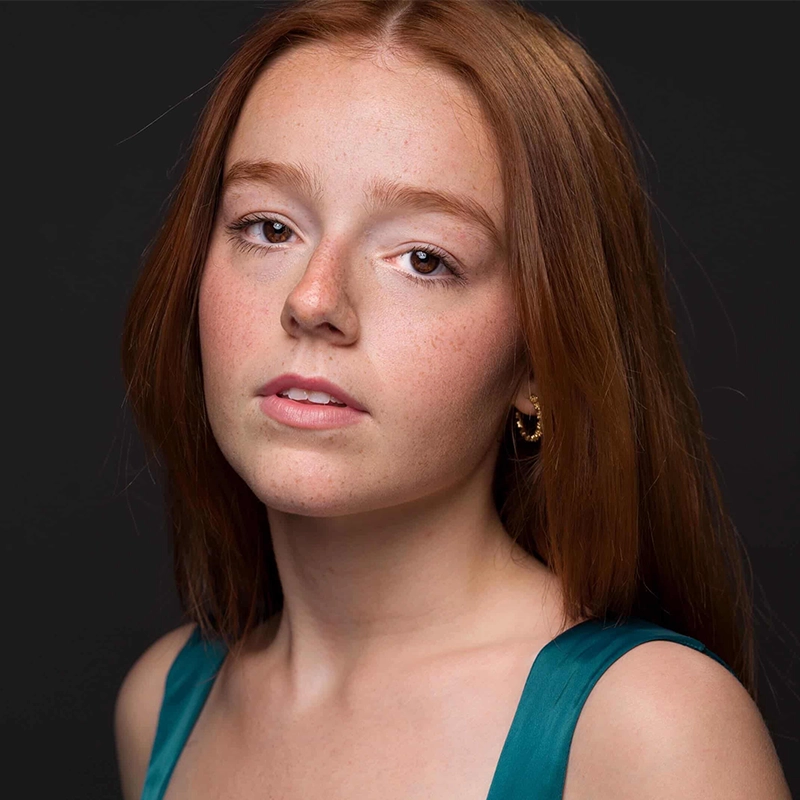 A young woman with long red hair and fair, freckled skin gazes at the camera. Reminiscent of Millie Yocum, she wears a teal sleeveless top and small gold hoop earrings. The dark background enhances her features and gentle expression, creating a striking visual Surfac.