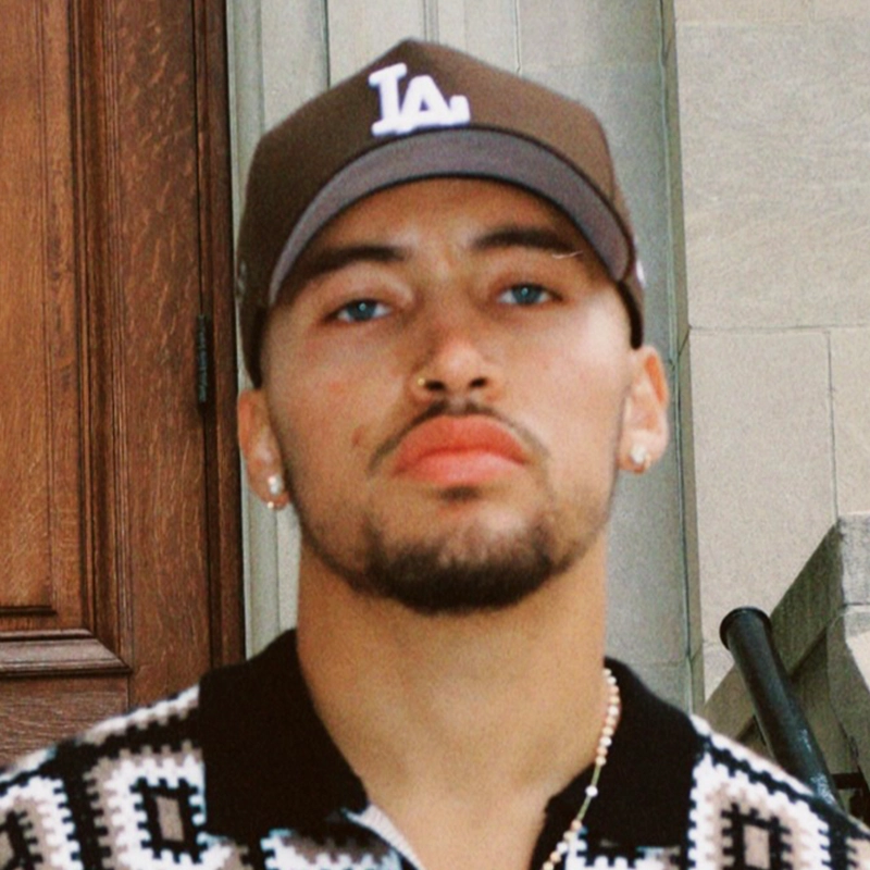 A young man with a trimmed beard and mustache wears a brown LA baseball cap, stud earrings, and a patterned polo shirt. He has piercing blue eyes and a serious expression, standing in front of a wooden door and stone wall.