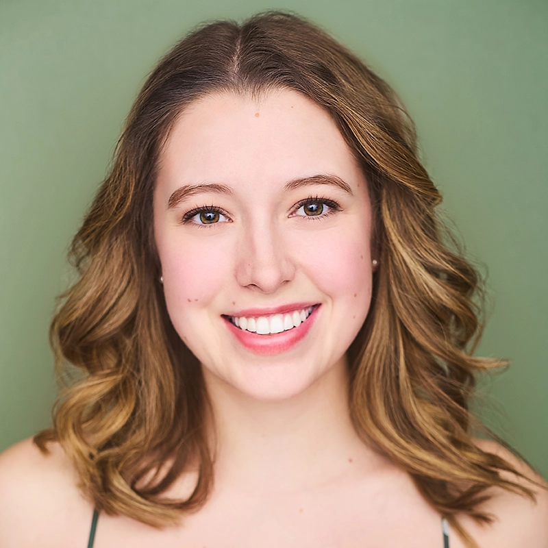 A young woman, Casey Keeler, with wavy light brown hair smiles at the camera. She has fair skin, blue eyes, and wears a green top. The background is a solid soft green, giving the portrait a warm, inviting feel. The lighting is even and highlights her features.