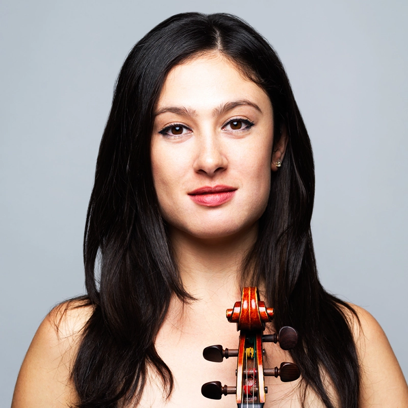 Gabby Sherba, a woman with long dark hair, stands against a gray background, holding a violin upright in front of her. She has light skin, dark eyes, and a neutral expression. The focus is on her face and the violin’s scroll, which is visible in the foreground.