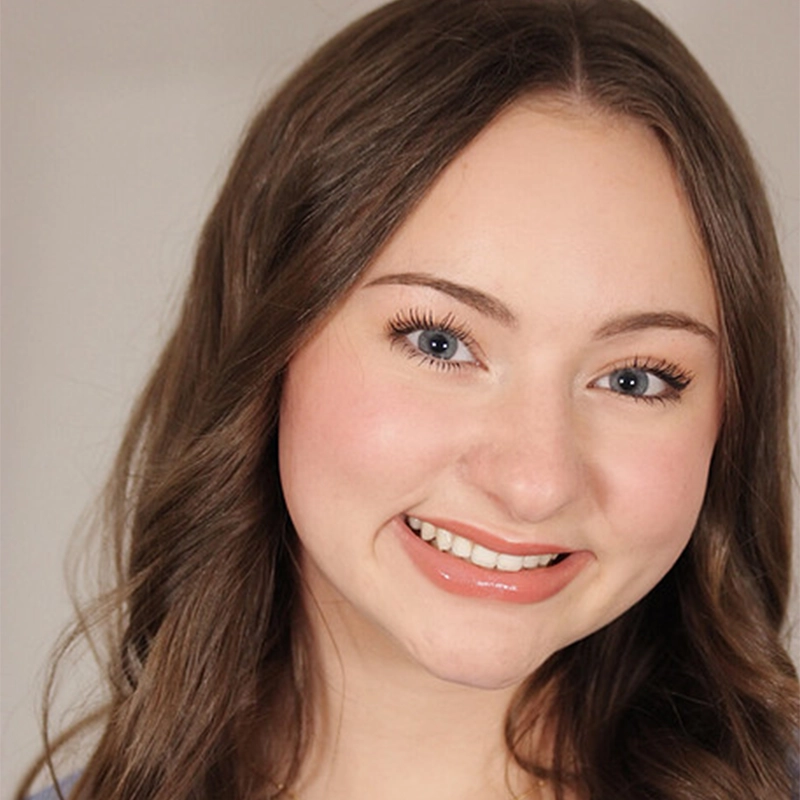 Young woman with long, wavy brown hair and blue eyes smiles at the camera. She has natural makeup, including pink blush and glossy nude lipstick. The background is a neutral light beige, and the lighting is soft and even.